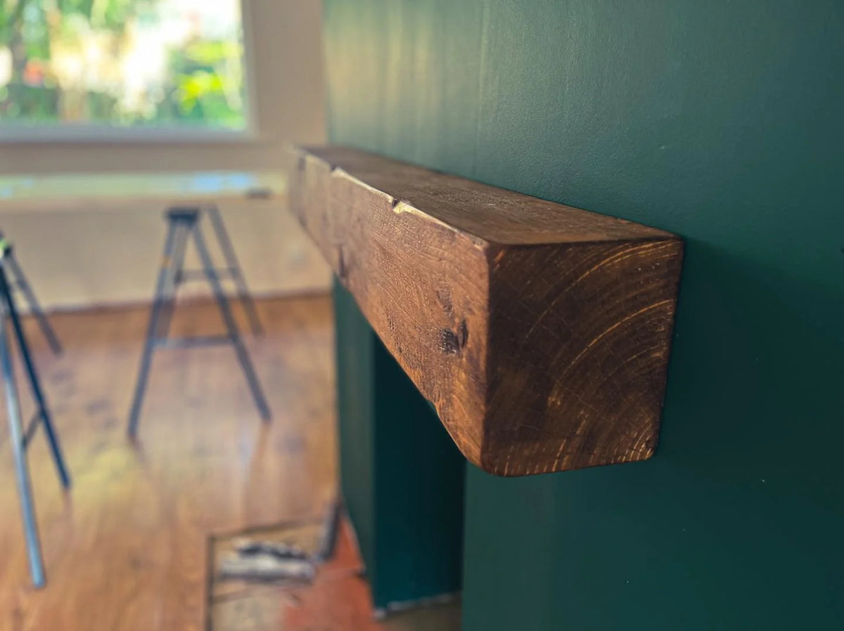 A rustic dark brown wooden mantel shelf mounted on a dark green wall in a home setting with a blurred window and sawhorses in the background.