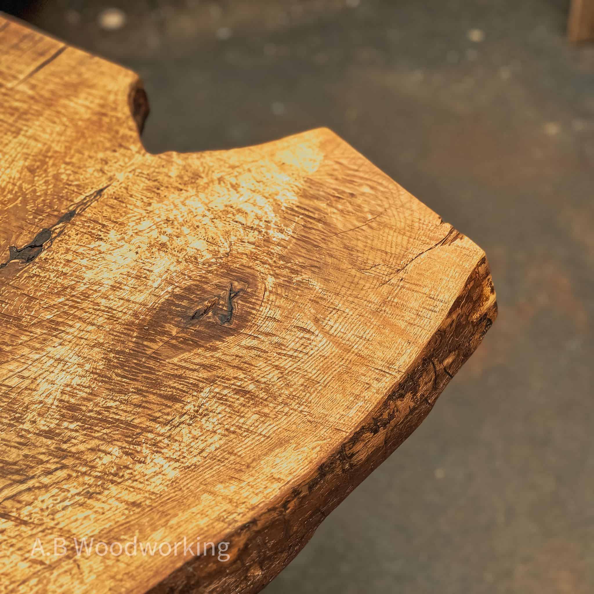 Close-up of a rustic live-edge wooden tabletop showing natural wood grain, knots, and rough bark edges, with a blurred workshop floor in the background.