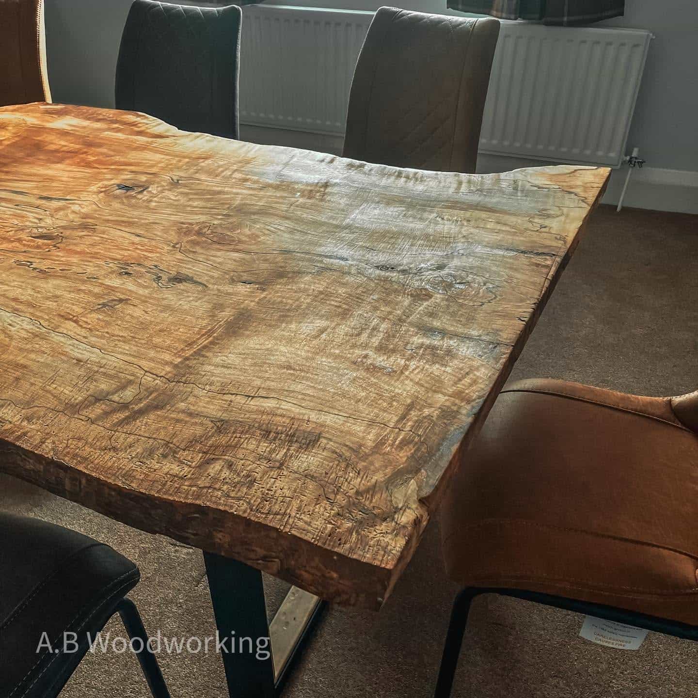A rustic live-edge wooden dining table with a rich natural grain, shown in a dining room setting with leather and upholstered chairs. A watermark for 'A.