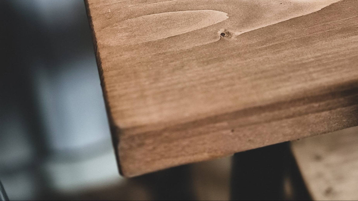 Close-up of a rustic wooden tabletop showing natural wood grain, knots, and a warm medium-brown finish, with a blurred dark support structure on the left.