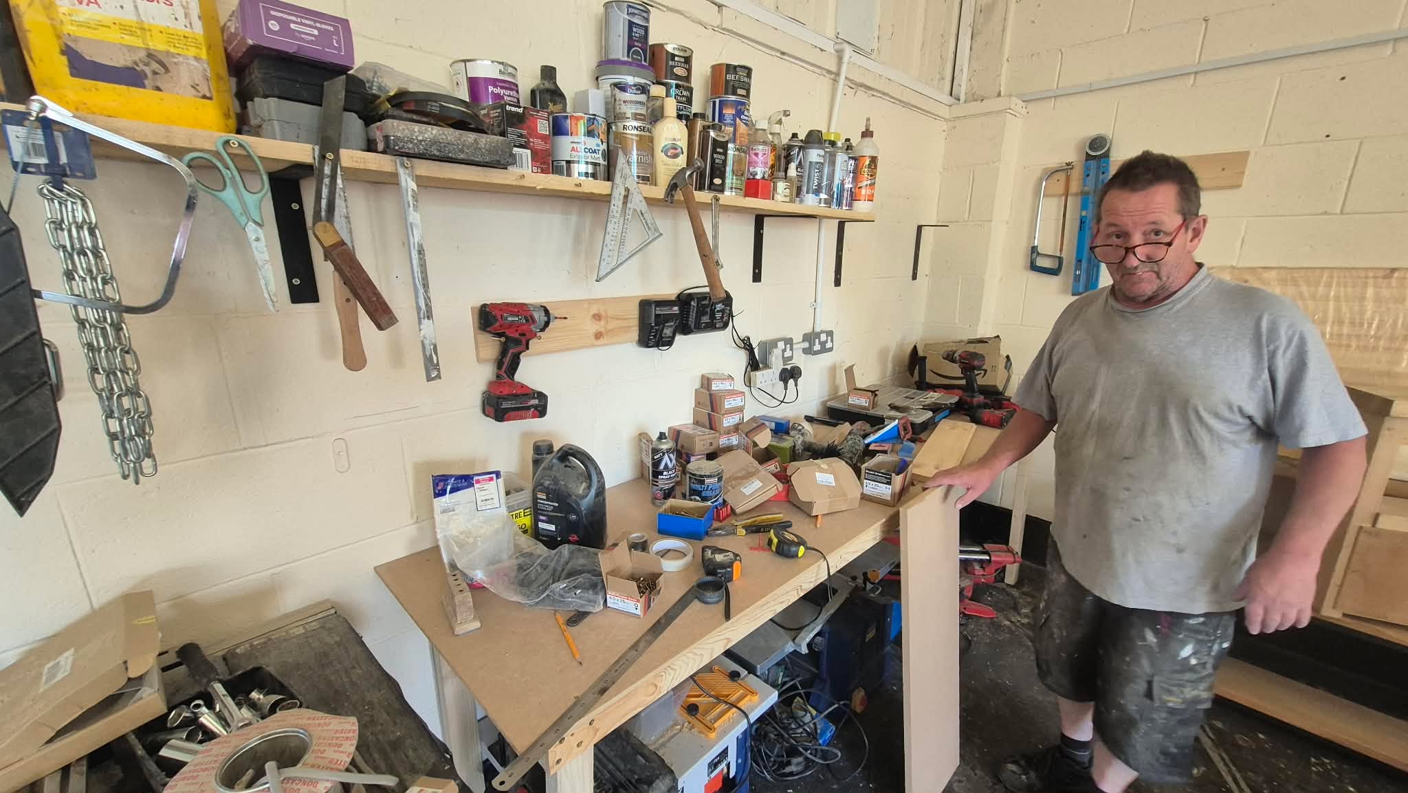 A man in a gray t-shirt and paint-splattered shorts stands in a cluttered workshop holding a piece of wood, surrounded by tools, shelves, and workbench clutter.